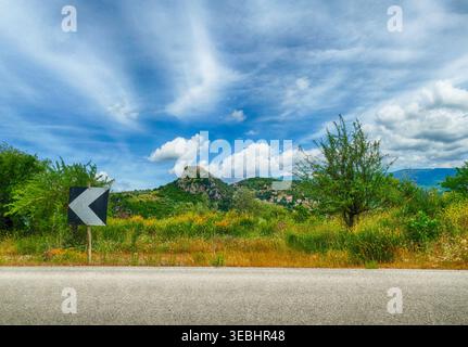 Route de Grèce menant à travers un paysage d'été près d'Olympia, avec un panneau de chevron sur le bord de la route, des collines verdoyantes, des fleurs sauvages, des montagnes, et un blu Banque D'Images