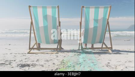 Assis couple mature portant des vêtements de plage décontractés se relaxant sur la plage de sable, avec chaises longues rayées Banque D'Images