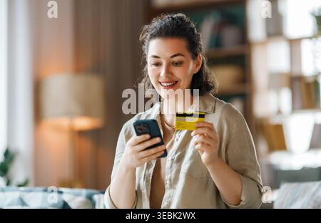 Portrait d'une femme joyeuse assise dans un salon confortable, utilisant son smartphone et tenant une carte de crédit. Elle a l'air excitée et satisfaite, highlightin Banque D'Images