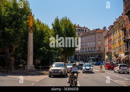 Trieste, Italie - 12 août 2025 : une scène de rue animée à Trieste, Italie, avec une statue religieuse dorée éminente au sommet d'une haute colonne. Le comp Banque D'Images