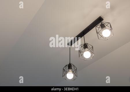 Luminaire de plafond industriel moderne suspendu au plafond lumineux. Suspension géométrique en métal noir diffusant une lueur invitante à travers l'intérieur de la pièce blanche, cr Banque D'Images