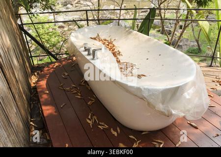 Baignoire extérieure blanche reposant sur une terrasse en bois avec couverture en plastique collectant les débris de feuilles d'automne sèches dans un cadre paisible et calme entouré d'arbres Banque D'Images