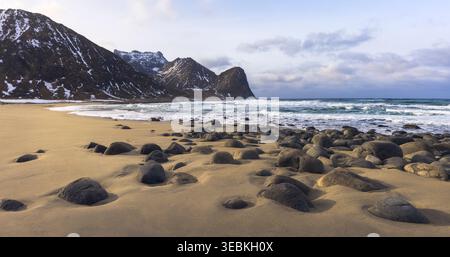 Unstad Beach présente de puissantes vagues qui s'écrasent sur des rochers lisses, avec des montagnes majestueuses derrière, créant une scène côtière sereine, Lofoten, Norvège Banque D'Images