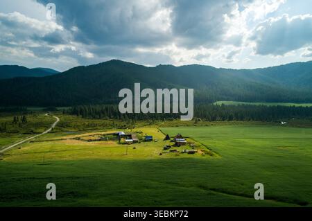 La vue aérienne capture de vastes champs verts entourant une propriété isolée au pied d'imposantes montagnes boisées sous un ciel nuageux Banque D'Images