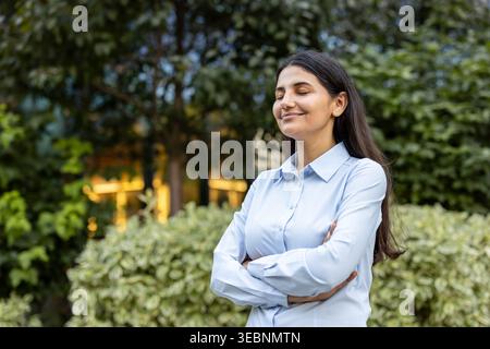 Jeune femme d'affaires en plein air avec les bras croisés, les yeux fermés et souriante, profitant d'un moment paisible de calme et d'équilibre pendant une journée de travail chargée Banque D'Images