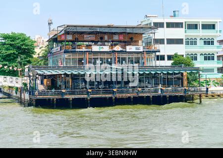 BANGKOK, THAÏLANDE - 27 février 2025 : Café au bord de la rivière sur pilotis au-dessus de la rivière Chao Phraya à Bangkok avec de grandes fenêtres en verre et une terrasse ouverte. Banque D'Images