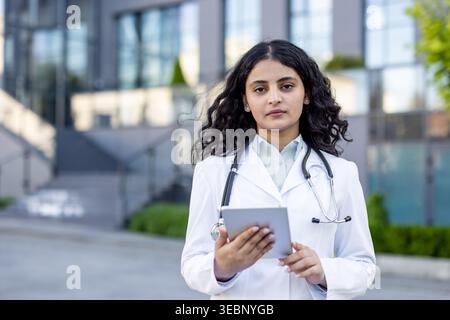 Jeune médecin indienne portant une blouse de laboratoire blanche et un stéthoscope, tenant une tablette numérique tout en se tenant en toute confiance devant un bâtiment hospitalier contemporain Banque D'Images