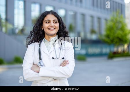 Jeune femme médecin avec des cheveux bouclés foncés et un stéthoscope autour du cou souriant avec confiance avec les bras croisés, tout en se tenant dehors devant un bâtiment moderne Banque D'Images