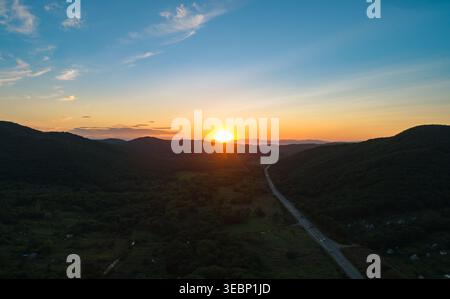 La vue aérienne capture un coucher de soleil vibrant descend sur des montagnes verdoyantes et une route sinueuse dans une vallée paisible au crépuscule Banque D'Images