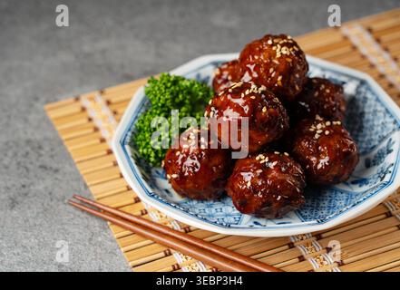 Délicieuses boulettes de viande glacées avec sauce aigre-douce, graines de sésame et baguettes sur un tapis de bambou Banque D'Images