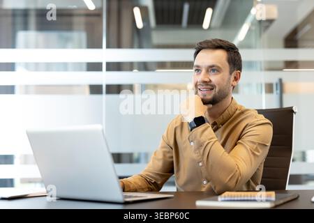Homme d'affaires assis au bureau avec ordinateur portable, souriant et regardant loin, contemplant les idées et opportunités futures d'affaires, représentant l'ambition et le succès Banque D'Images