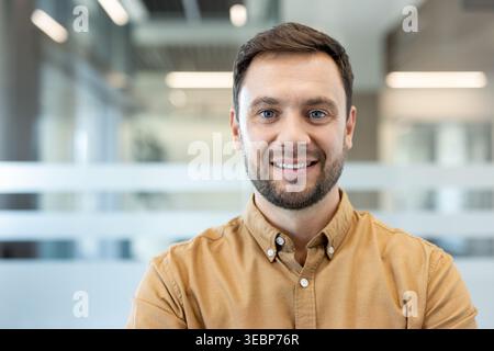 Jeune homme d'affaires professionnel avec la barbe et les yeux bleus debout avec confiance dans un bureau moderne lumineux, souriant et projetant le leadership, l'ambition et le succès dans un cadre d'entreprise Banque D'Images