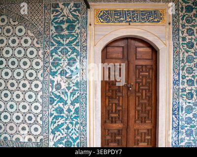Porte ancienne en bois avec des sculptures géométriques complexes serties dans un mur de carreaux de céramique iznik bleus et blancs ornés et calligraphie arabe dorée Banque D'Images