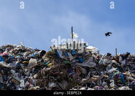 Un site d'enfouissement montre un grand tas de déchets avec divers articles dispersés à travers. Un oiseau vole au-dessus des déchets sous un ciel bleu clair. Banque D'Images