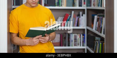 Jeune homme en chemise jaune lit livre tout en se tenant debout dans le cadre confortable de la bibliothèque Banque D'Images