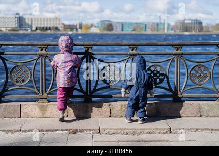 Les tout-petits portant des vêtements saisonniers à capuche regardent l'eau à travers une clôture de fer ornée sur une promenade pavée en Lettonie avec un architecte urbain moderne Banque D'Images