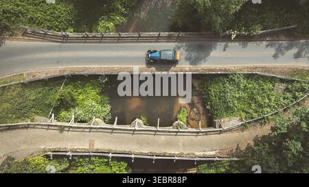 Une photo au-dessus du ciel capture un véhicule d'époque traversant un pont de pierre, entouré d'une végétation luxuriante et d'une rivière qui coule Banque D'Images