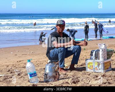 Jeune homme assis sur la plage avec des rafraîchissements à vendre à Anchor point, Taghazout, Maroc, avec des surfeurs se préparant en arrière-plan. Banque D'Images