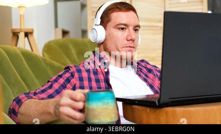 Vue à hauteur des yeux d'un homme portant des écouteurs blancs, tenant une tablette et une tasse à café dans un salon confortable. Lecture numérique, télétravail et audio Banque D'Images