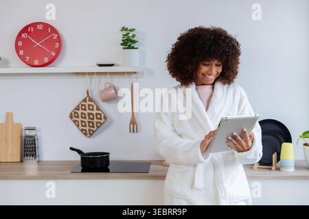 Femme souriante naviguant sur Internet sur tablette dans la cuisine portant un peignoir Banque D'Images