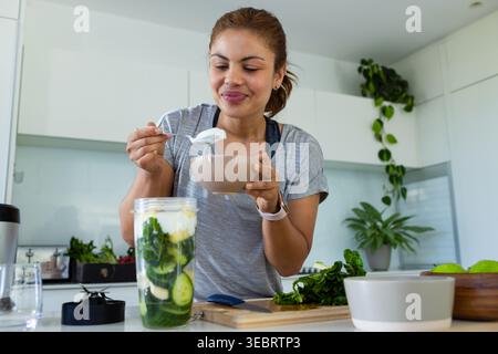 Femme africaine adulte tenant un bol beige et une cuillère tout en mélangeant un smoothie vert à l'îlot de cuisine Banque D'Images