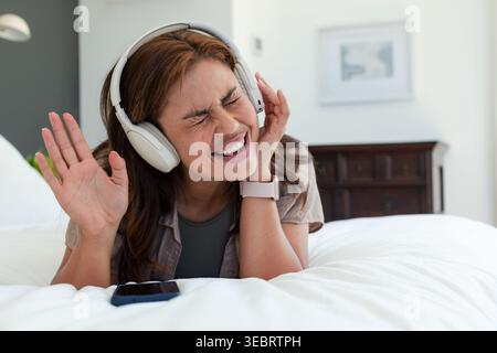 Femme afro-américaine couchée sur un lit blanc chantant, touchant un casque blanc, smartphone à proximité Banque D'Images