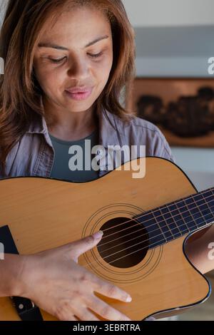 Guitariste féminine tenant une guitare acoustique, plumant des cordes et regardant le fretboard à la maison Banque D'Images