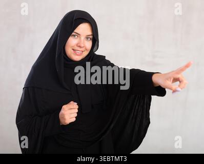 Joyeuse jeune femme musulmane vêtue de hijab noir pointant sur le côté Banque D'Images