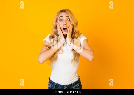 Surprise jeune femme exprimant son étonnement avec les mains sur son visage devant un fond jaune Banque D'Images