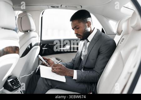 Homme d'affaires travaillant sur un bloc-notes dans le siège arrière d'une voiture de luxe pendant une journée bien remplie Banque D'Images
