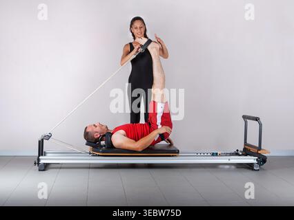 instructeur féminin en vêtements sportifs noirs guide un homme en chemise rouge alors qu'il effectue un étirement de jambe en utilisant les sangles d'un réformateur de pilates professionnel Banque D'Images