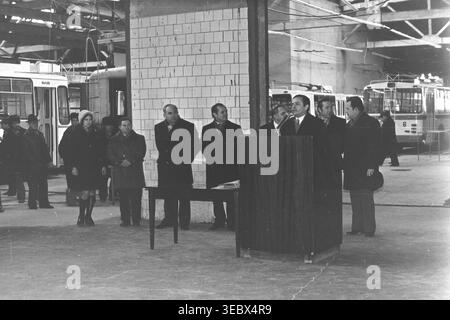 Sloviansk, 1986. Une réunion solennelle sur le territoire du dépôt de trolleybus. Un fonctionnaire en manteau prononce un discours depuis un podium en bois, entouré de membres de l'administration et de la direction. L'événement marque une étape importante dans le développement des infrastructures de transport de la ville. Les travailleurs se rassemblent pour écouter l'adresse dans le spacieux garage de l'entreprise. Cette photographie historique d'archives capture l'atmosphère sociale et de travail d'une entreprise municipale soviétique, préservant la mémoire d'un Donbass pacifique. Banque D'Images