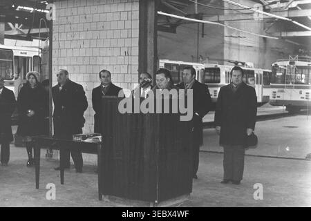 Sloviansk, 1986. Une réunion solennelle sur le territoire du dépôt de trolleybus. Un fonctionnaire en manteau prononce un discours depuis un podium en bois, entouré de membres de l'administration et de la direction. L'événement marque une étape importante dans le développement des infrastructures de transport de la ville. Les travailleurs se rassemblent pour écouter l'adresse dans le spacieux garage de l'entreprise. Cette photographie historique d'archives capture l'atmosphère sociale et de travail d'une entreprise municipale soviétique, préservant la mémoire d'un Donbass pacifique. Banque D'Images