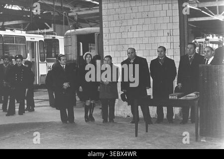 Sloviansk, 1986. Une réunion solennelle sur le territoire du dépôt de trolleybus. Un fonctionnaire en manteau prononce un discours depuis un podium en bois, entouré de membres de l'administration et de la direction. L'événement marque une étape importante dans le développement des infrastructures de transport de la ville. Les travailleurs se rassemblent pour écouter l'adresse dans le spacieux garage de l'entreprise. Cette photographie historique d'archives capture l'atmosphère sociale et de travail d'une entreprise municipale soviétique, préservant la mémoire d'un Donbass pacifique. Banque D'Images