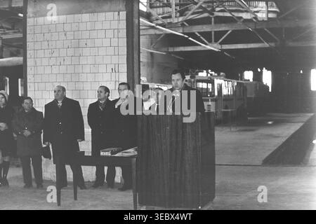 Sloviansk, 1986. Une réunion solennelle sur le territoire du dépôt de trolleybus. Un fonctionnaire en manteau prononce un discours depuis un podium en bois, entouré de membres de l'administration et de la direction. L'événement marque une étape importante dans le développement des infrastructures de transport de la ville. Les travailleurs se rassemblent pour écouter l'adresse dans le spacieux garage de l'entreprise. Cette photographie historique d'archives capture l'atmosphère sociale et de travail d'une entreprise municipale soviétique, préservant la mémoire d'un Donbass pacifique. Banque D'Images