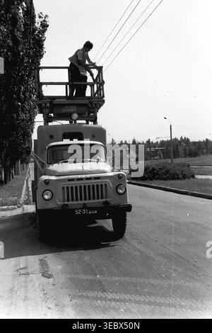 Sloviansk, 1986. Deux techniciens effectuent l'entretien du réseau aérien de contact du système de trolleybus de la ville. Les ouvriers sont élevés dans le panier d’un camion utilitaire gaz spécialisé garé sur le côté d’une large route urbaine. Portant des casques de sécurité, ils inspectent et réparent soigneusement les lignes électriques assurant le fonctionnement fiable des transports publics. Cette photographie documentaire historique met en évidence le travail industriel quotidien et l'entretien des infrastructures de la période soviétique, capturant l'environnement urbain d'un Donbass paisible. Banque D'Images