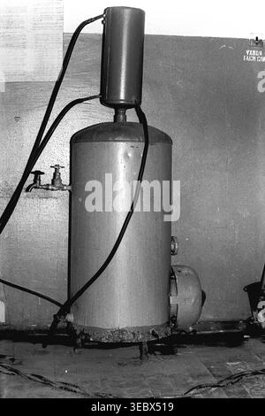 Sloviansk, 1986. Une grande chaudière utilitaire de chauffage de l'eau installée à l'intérieur des locaux utilitaires du dépôt de trolleybus. Le réservoir métallique cylindrique, montrant des signes d'usure et de corrosion à sa base, est relié à des tuyaux, des vannes et un vase d'expansion plus petit au-dessus. Cet équipement fournissait de l'eau chaude ou du chauffage essentiels aux travailleurs de l'installation et aux ateliers de réparation. Cette photographie d'archives en noir et blanc offre un aperçu brut de l'infrastructure industrielle quotidienne et de l'ingénierie pragmatique de l'ère soviétique, préservant un Donbass paisible. Banque D'Images