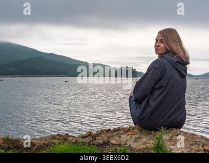 Jeune femme en sweat à capuche gris se relaxant par l'eau tranquille avec des montagnes vertes et une petite île sous le ciel couvert Banque D'Images