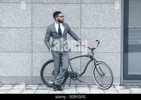 Homme d'affaires en costume debout à côté de vélo contre le bâtiment moderne pendant la journée Banque D'Images