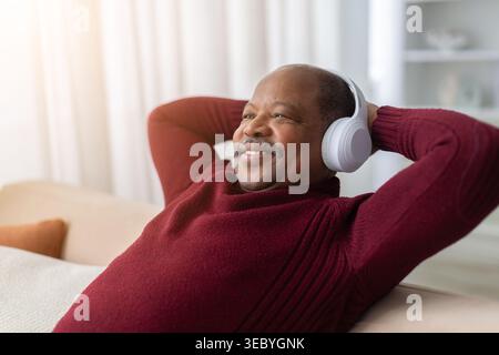 Homme afro-américain mature détendu allongé sur le canapé portant des écouteurs sans fil Banque D'Images