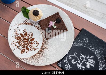 Un dessert au brownie végétalien plaqué servi sur une assiette blanche texturée, accompagné d’une boule de glace végétalienne garnie d’un biscuit et de menthe fraîche. Banque D'Images