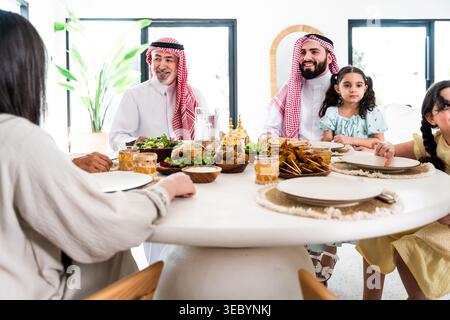 Famille arabe multigénérationnelle réunie autour d'une table à manger dans leur maison moderne, partageant repas traditionnel, conversation et hospitalité chaleureuse pendant Banque D'Images