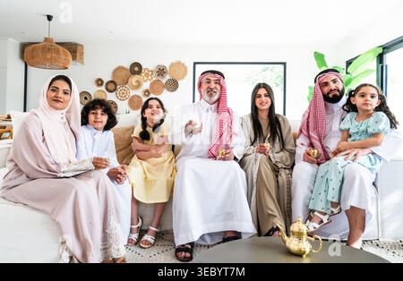 Famille arabe multigénérationnelle assis sur un canapé dans leur salon moderne, les parents et les enfants appréciant le temps de qualité tout en regardant la télévision et Banque D'Images