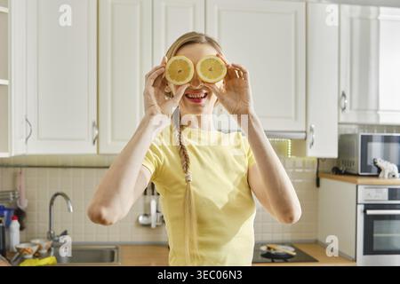 Femme ludique se tient dans une cuisine tenant deux tranches de citron devant son visage. Elle est souriante et profite de son temps tout en préparant un repas. Le k Banque D'Images