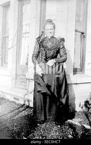 Cette photographie en noir et blanc capture une femme victorienne âgée debout à l'extérieur devant un bâtiment en pierre ou une maison avec de grandes fenêtres à guillotine. Elle porte une robe sombre et formelle avec un corsage à col haut orné de motifs ou de dentelle avec des épaules structurées, et ses cheveux sont soigneusement attachés en arrière dans un chignon. Extrait d'un album victorien tardif de petites photographies prises par un photographe amateur. La plupart datent de 1898 à 1901 et semblent se rapporter à la famille de John White (peut-être de Sth London), un constructeur et entrepreneur. La qualité des photographies originales était variable Banque D'Images