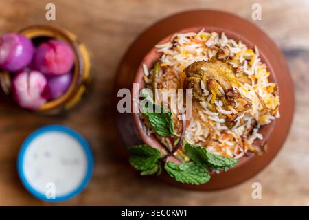 Vue rapprochée du ciel d'un biryani de poulet savoureux dans un pot traditionnel en argile, accompagné d'oignons marinés et d'une sauce raita crémeuse au yaourt. Banque D'Images