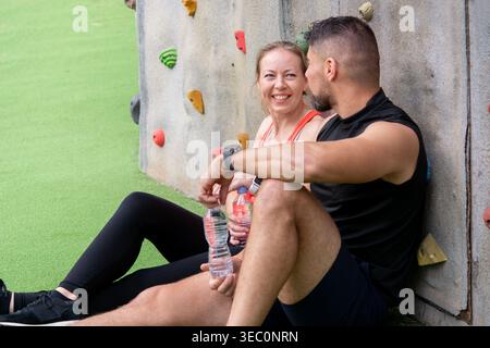 Heureux couple athlétique assis et relaxant après l'entraînement en plein air. Souriant homme et femme se reposant près du mur d'escalade, l'eau potable et jouissant Activ Banque D'Images