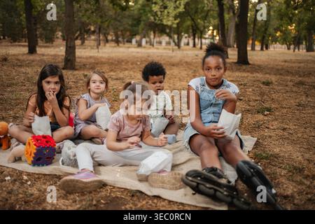 Cinq enfants sont assis sur une couverture au parc, mangeant des collations et regardant un film Banque D'Images