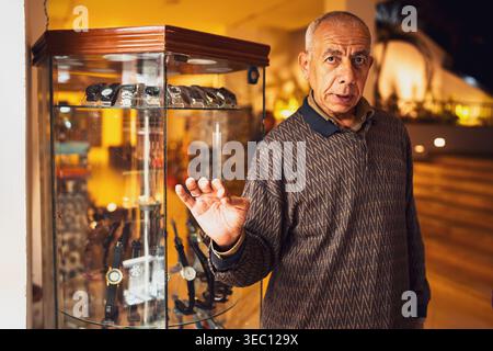 Marsa alam, égypte 21 avril 2026 : un horloger égyptien âgé à marsa alam se tient près de sa vitrine de montres, souriant dans la lumière chaude du soir Banque D'Images