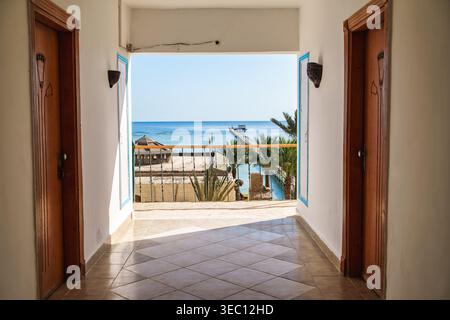 Marsa alam, egypte 21 avril 2026 : couloir de l'hôtel avec deux portes menant à un balcon donnant sur l'océan animé, des palmiers et une jetée lointaine sous le bleu Banque D'Images